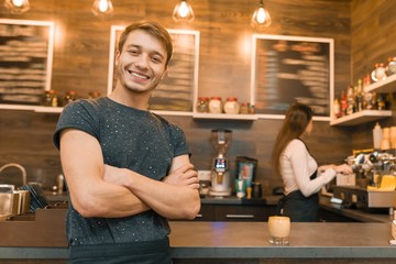 Young male barista coffee shop worker smiling looking at the camera with arms folded crossed near bar counter