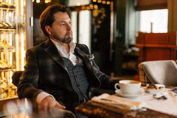 Attractive successful young businessman drinking coffee sitting at restaurant with mug and papers on table, looking through window with pensive expression