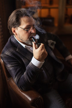 Pensive mature bearded businessman in expensive stylish suit and white shirt, no tie, smoking pipe in dark interior with dim lighting, thinking about forthcoming important meeting with partners