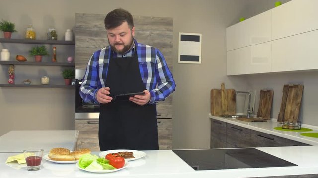 Fat caucasian man with a beard in an apron and with a tablet checks the ingredients for making burger, fast food