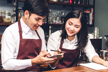 Portrait of couple small business owner smiling and working behind the counter bar in a cafe.Barista  using smartphone and receive order from customer