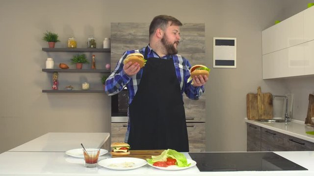 Satisfied fat man with a beard and apron dancing and smiling in the kitchen with burgers