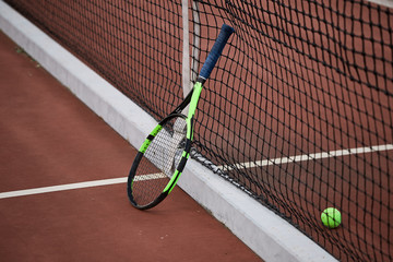 Single tennis racquet lying with a yellow ball on a red-clay tennis court background. Tennis is a...