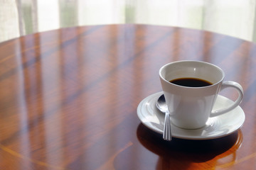 Closeup of white glass of black coffee with saucer and metal spoon on glossy wooden round table and curtain background of window