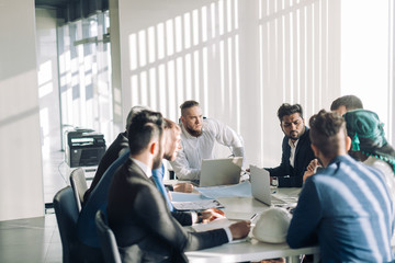 Smiling confident executive in white shirt sitting with laptop at meeting table with business partners team, discussing the working issues.