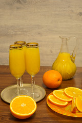 Three glasses of mimosa cocktail, a decanter with orange juice and fresh oranges on a wooden table