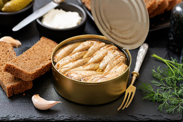 Canned sprats in tin can with bread for making sandwich on dark  background.