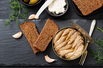 Canned sprats in tin can with bread for making sandwich on dark  background.