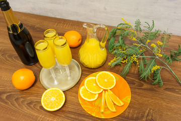 Three glasses of mimosa cocktail, a bottle of champagne, a decanter with orange juice, fresh oranges and mimosa sprig on a wooden table