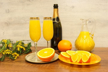 Two glasses of mimosa cocktail, a bottle of champagne, a decanter with orange juice, fresh oranges and mimosa sprig on a wooden table