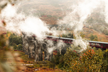 Fototapeta na wymiar Panorama of red steam train passing Glenfinnan Viaduct with fog covered mountains during a moody autumn day (Glenfinnan, Scotland, Europe)