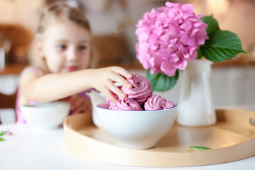 Cute kid girl takes pink marshmallows from table. Children tea time in kitchen. White mug, sweets, cupcakes and hydrangea flowers are serving on wooden tray. Cozy home atmosphere hygge.