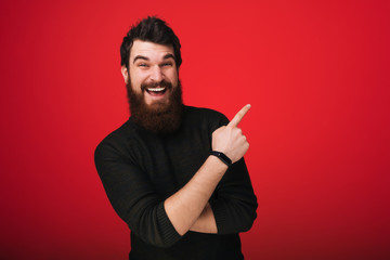 Fototapeta premium Good-looking bearded man in casual sweater pointing right with index finger and smiling cheerfully