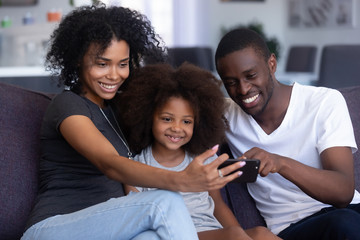 Happy african family with child daughter laugh have fun with gadget on sofa look at smartphone take selfie at home, black parents and kid watch funny video, make online call on phone, use mobile app