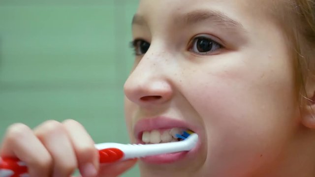 Close-up cute beautiful female child with fair hair brushes her teeth thoroughly and smiles in bathroom on background wall from green tile