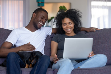 Smiling african american couple laughing using computer sitting on couch with pet dog, happy black...