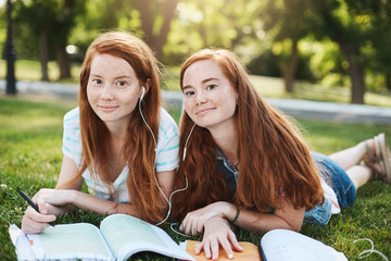 Girls make everything together. Charming natural redhead women in summer clothes lying on grass during weekends, sharing earphones to listen songs together, sister trying help with homework