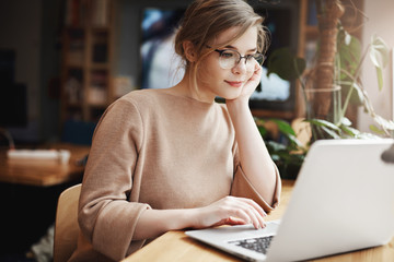 Girl gazing at profile page of handsome man, browsing through dating sire while sitting in cafe with laptop, leaning on hand and gazing at notebook screen, sitting in cafeteria near window