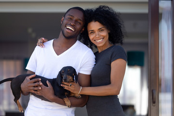 Happy african american millennial couple embrace outdoors holding dachshund, smiling young black man woman enjoying time outside house having fun with dog looking at camera, family and pet portrait