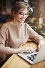 Vertical shot of successful and happy european female student in trendy glasses and casual outfit, smiling broadly while sitting near window in cafe and using laptop to update social page
