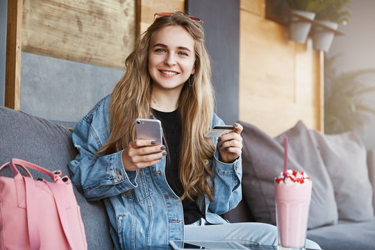 Good-looking fair-haired girl in trendy denim jacket sitting and relaxing in cafe, holding smartphone and credit card, wanting to pay for delicious cocktail, smiling cheerfully at camera