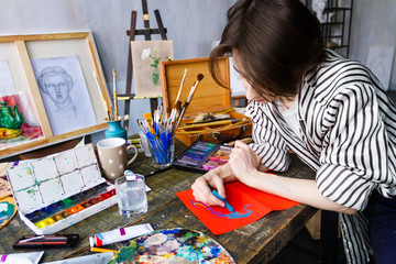 Pensive woman artist is drawing sketch with pastels on orange sheet of paper in art studio. Creative workplace on table with drawing tools, brushes, pastels, tubes, watercolors, palette. Art concept.