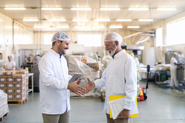 Two businessmen in sterile uniforms shaking hands for good done job while standing in food factory. Older one holding folder and graphs while younger holding tablet.