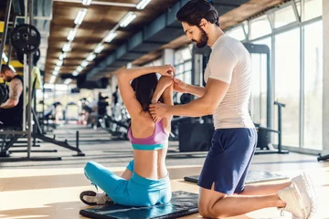 Poster Serious bearded personal trainer helping woman to stretch arms. Woman sitting on the mat, gym interior. © dusanpetkovic1