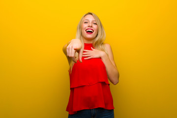 Young girl with red dress over yellow wall pointing with finger at someone and laughing a lot