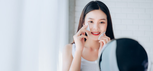 Young Beauty Asian Woman Cleaning Face with Cotton Her Looking at Mirror Clear Face Skincare and Smile Morning in White Bedroom,Copy Space Panoramic Web Banner.