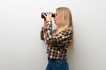 Blonde young girl over white wall and looking in the distance with binoculars
