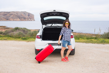 Travel, tourism and people concept - happy young woman going to travel by car with huge suitcase wearing glasses