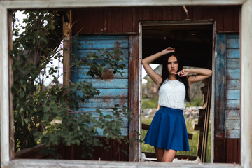 Young adult seductive brunette in blue skirt and white shirt posing in rustic house outdoors