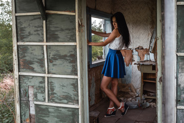 Young adult seductive brunette in blue skirt and white shirt posing in rustic house outdoors