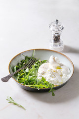 Sliced Italian burrata cheese, fresh arugula salad, pine nuts and olive oil in white ceramic plate on cloth over white marble table.