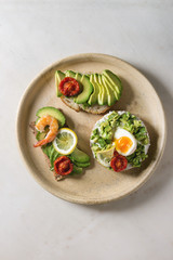Variety of vegetarian sandwiches with sliced avocado, sun dried tomatoes, egg, shrimps, arugula served on ceramic plate over white grey spotted background. Flat lay, space