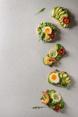 Fototapeta na wymiar Variety of vegetarian sandwiches with sliced avocado, sun dried tomatoes, egg, shrimps, arugula served over white grey spotted background. Flat lay, space
