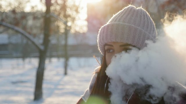 young pretty woman vaping on the street in the winter park at sunset