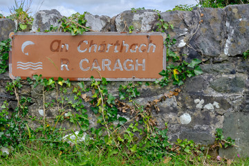 Sign on low road wall for River Caragh, both in English and in Gaelic.  Vine growing on wall and sign