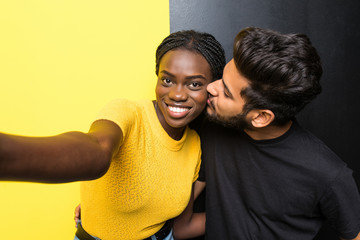 Young mixed race couple indian man kissing her african girlfriend in cheek standing isolated on yellow black background