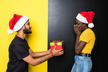 Young happy mixed race couple, indian man make a Christmas gift for his african woman wearing santa hats standing isolated on yellow black background
