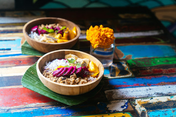 Close up smoothie bowl made with mango, banana, granola, grated coconut, dragon fruit, chia seeds and mint on colored background.