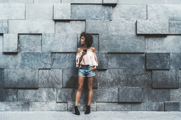 A dazzling young African-American female with a curly afro hair is leaning against the textured wall made of stone blocks with different displacement depth and typing a message via her smartphone