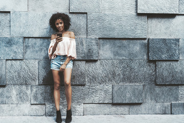 A flirtatious young mixed female with cellphone outdoors standing in front of a stone wall made with blocks of different displacement; with a copy space place on the right for your ad text or a logo