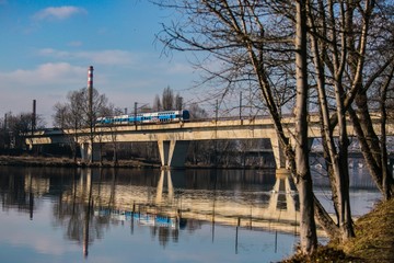 Prague, Czech Republic / Europe - January 31 2019: Railway bridge with train running across Moldau river at Liben, reflection in water, riverbank with trees, sunny day, blue sky