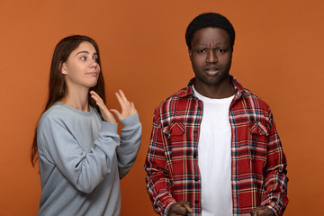 Studio image of angry young dark skinned male in red checkered shirt frowning, having displeased annoyed look quarreling with his white girlfriend who is feeling scared. Relationships and emotions