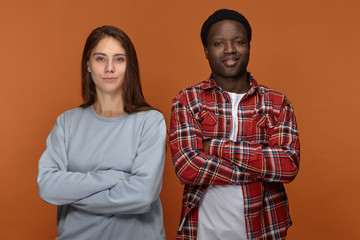 Horizontal shot of two young friends of different ethnicities standing next to each other. Attractive confident young dark skinned male posing in studio with his beautiful girlfriend, crossing arms