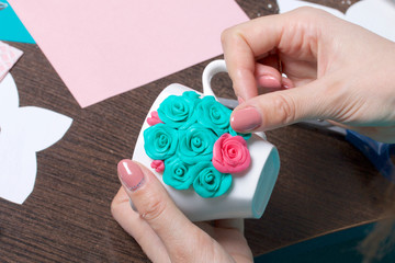 Crafts from polymer clay. A woman sticks a gummed polymer clay flower to a cup. Mug decorated with stucco made of polymer clay