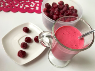 Cherry-banana smoothie poured into a glass. Nearby is a container of frozen cherries. Several berries are on the saucer.
