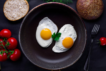 Fried eggs in a frying pan with dill, parsley, tomatoes cherry and sesame bun for a healthy breakfast. Top view. Protein food